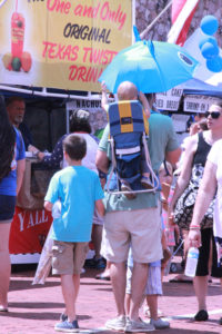 A father shields his baby from the sun with a unique blue umbrella and holds his son's hand as they enjoy the festival.